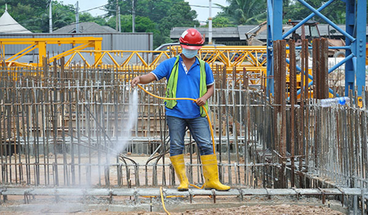 A construction worker, wearing safety gear, is spraying a rebar framework with water using a hose.
